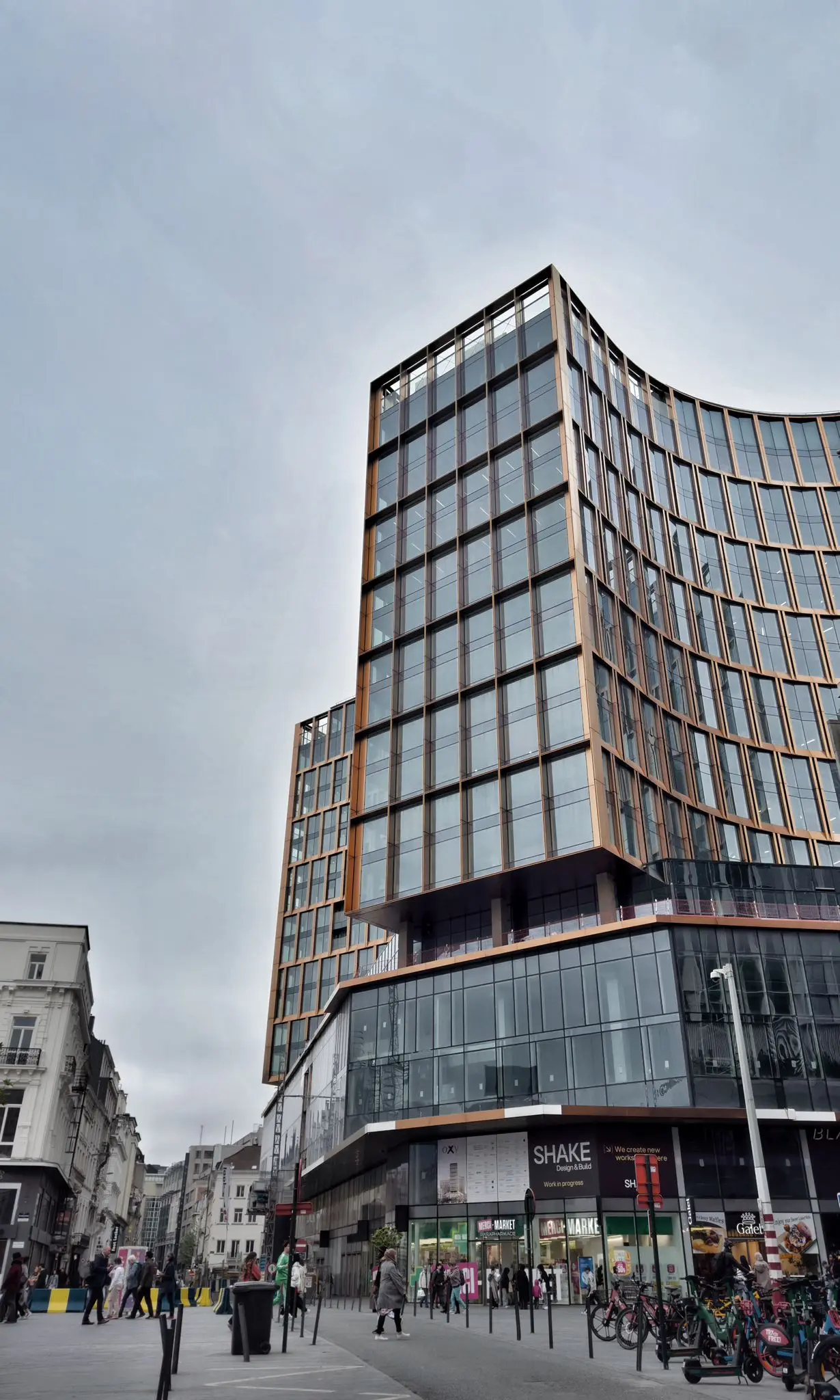 Modern, hoog glazen gebouw met afgeronde gevel op een stadsplein, met voorbijgangers, fietsen en winkels op het gelijkvloers.