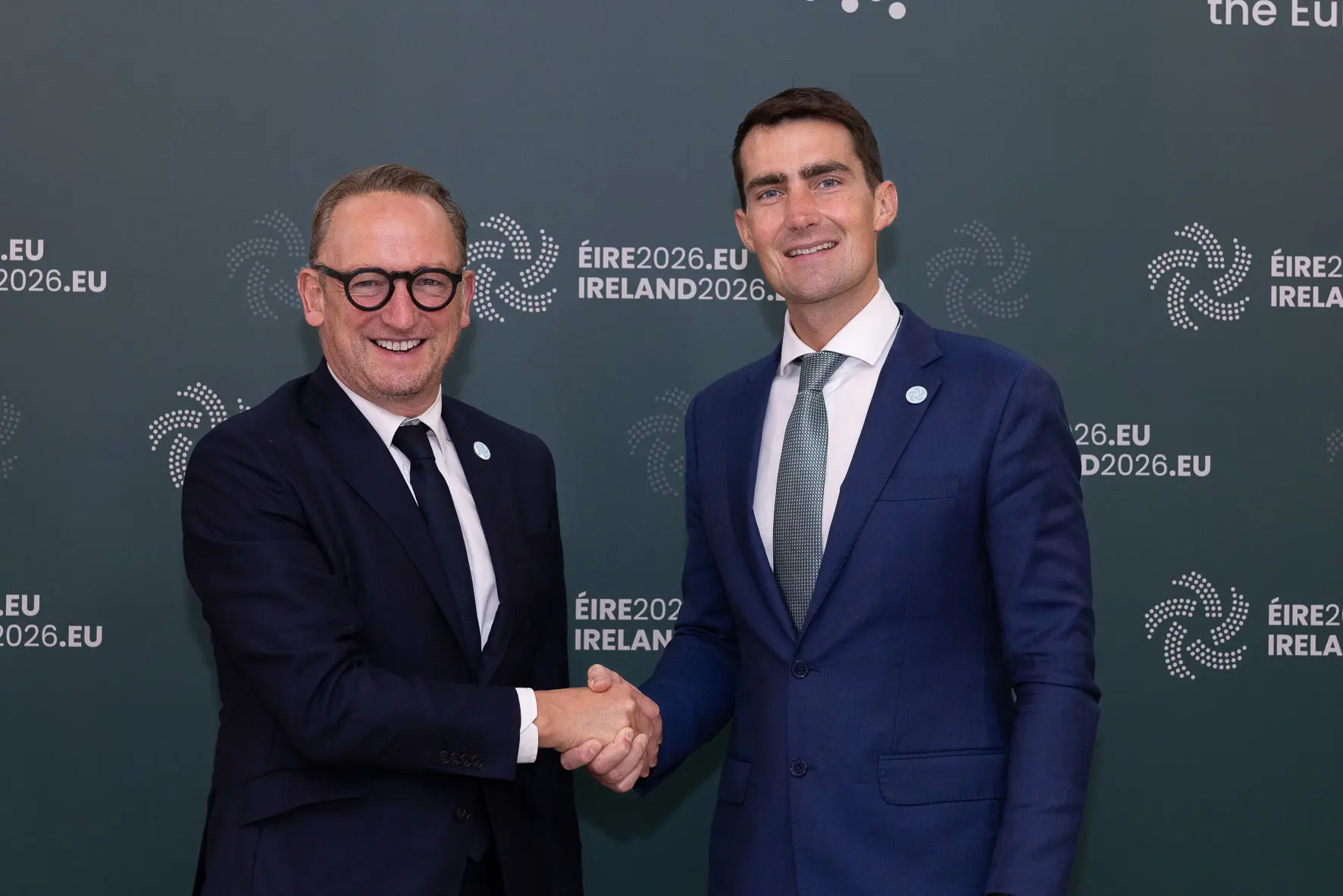 The two ministers shake hands in front of an Irish Presidency of the Council of the European Union backdrop.